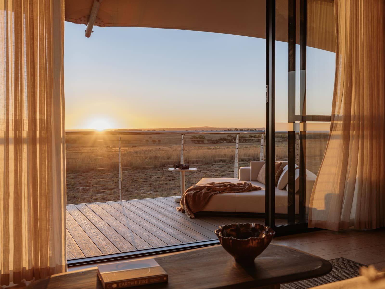 Sunrise over grassland, seen from a canvas lodge through open glass doors, with linen curtains and a daybed on the deck.