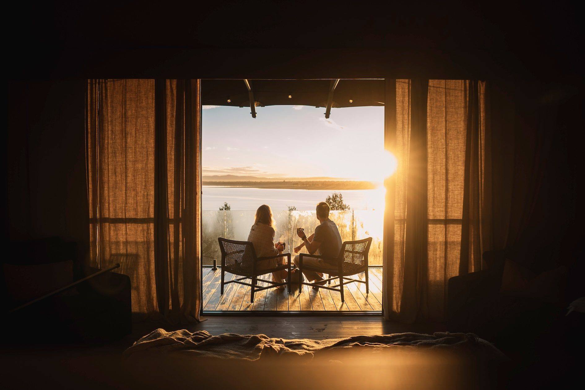 Two people seated in chairs on a timber deck at sunrise, seen from a dark room through linen curtains.