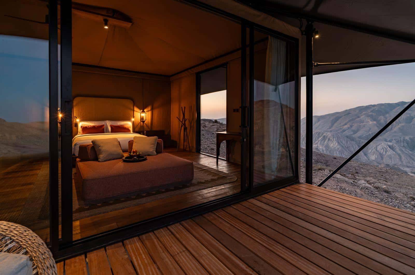Canvas bedroom with a low bed and rust-coloured cushions, opening through black-framed glass to a rocky mountain deck.