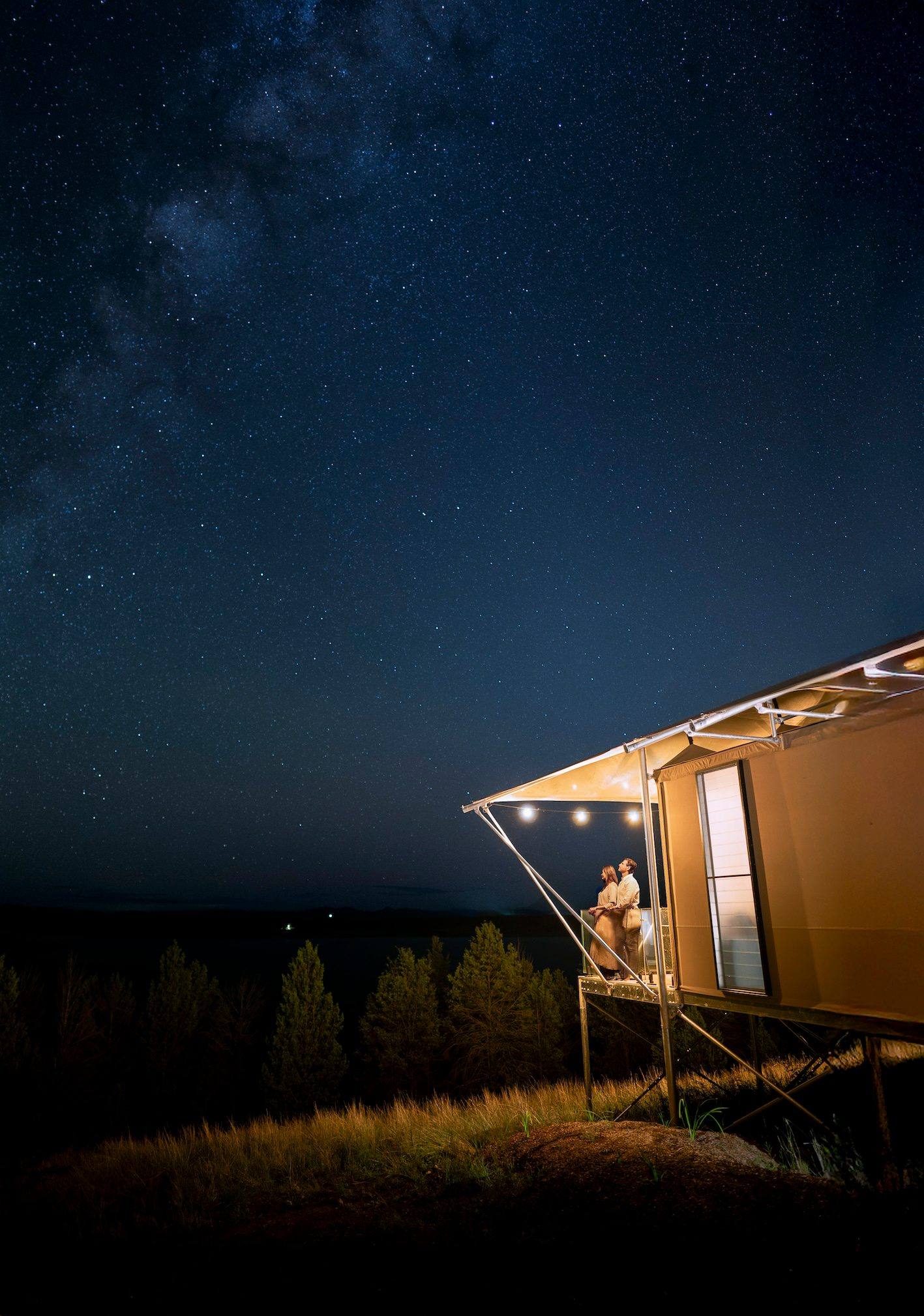 Canvas tent on a raised deck at night, with two people under a string of lights and the Milky Way overhead.