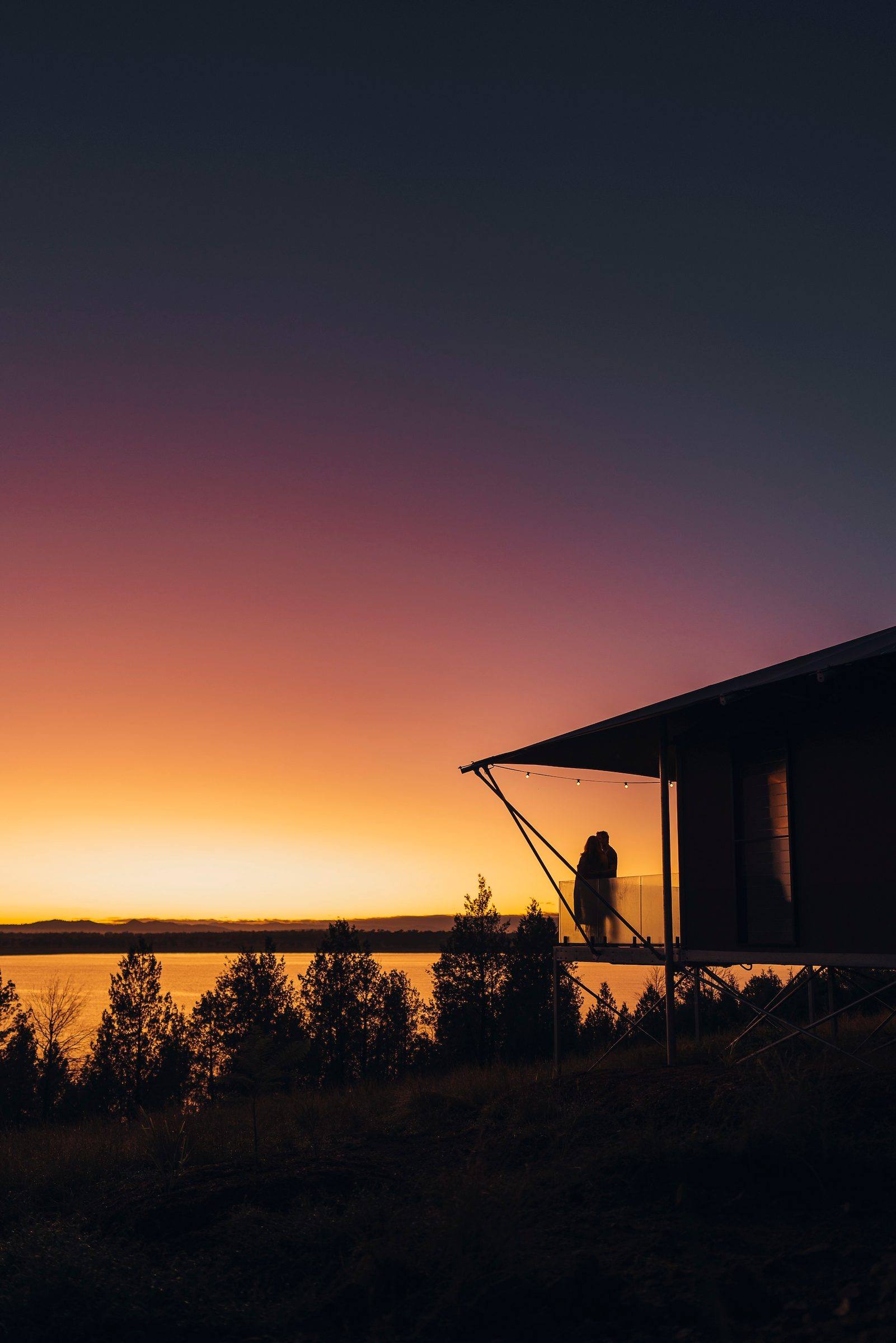 Two people silhouetted on the deck of a raised tent at sunrise, above pines and a lake.