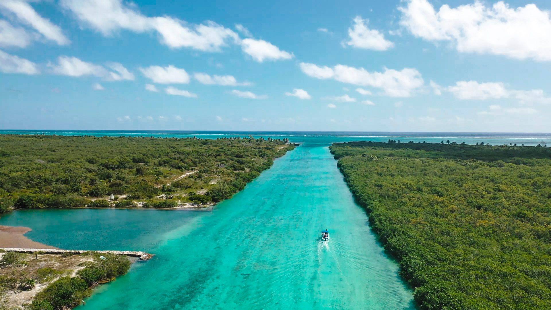 Aerial view of a turquoise channel through dense green forest, with a small boat on the water.