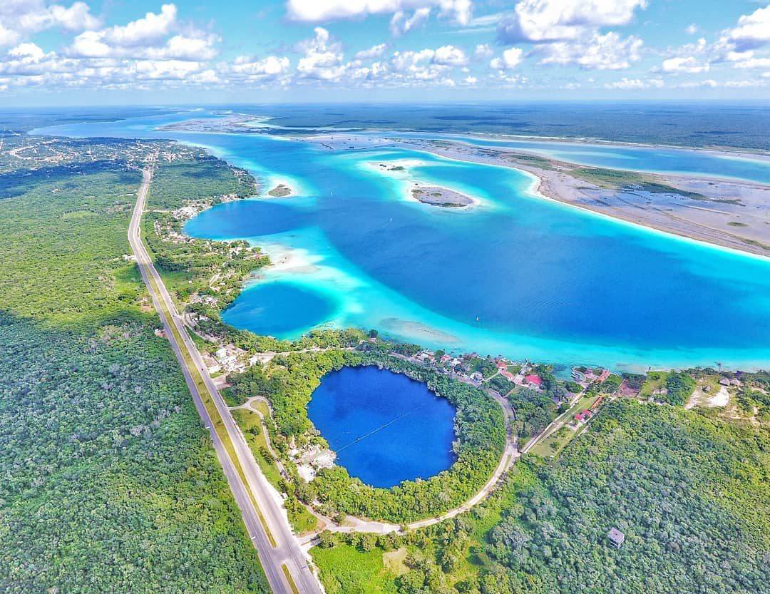 Aerial view of a turquoise lagoon, a round blue sinkhole, and forest beside a road.