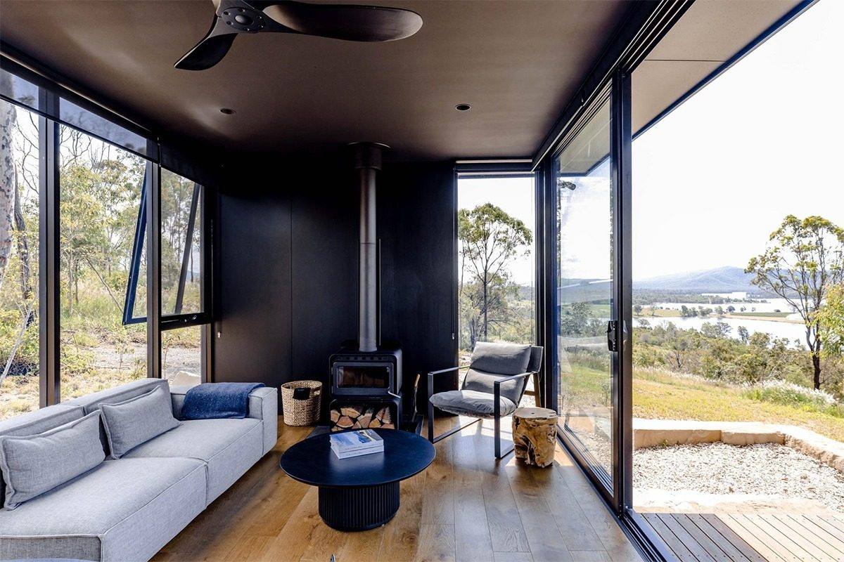 Living room with open awning windows, a wood stove, timber floor, and a view across hills to water.