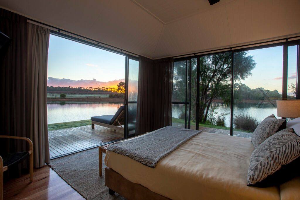 Bedroom with timber floor and linen bedding, opening through glass doors to a deck and a tree-lined lake.