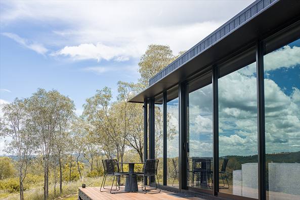 Timber deck and black-framed glass wall of a pavilion, with chairs facing pale-trunked trees.