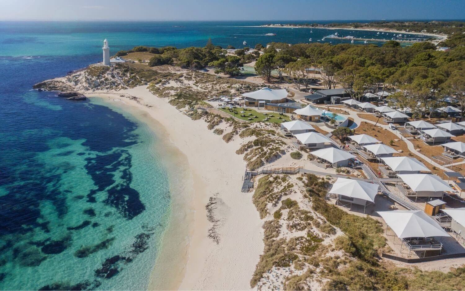 Aerial view of white canvas roofs along a sandy beach, with turquoise water and a white lighthouse on the headland.