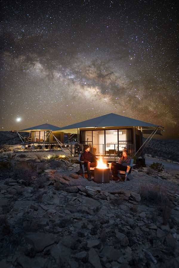 Two canvas-roofed lodges on rocky ground at night, with two people seated by a fire under the Milky Way.