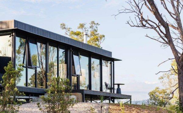 Dark-framed glass pavilion with open awning windows, raised above shrubs on a tree-lined slope.