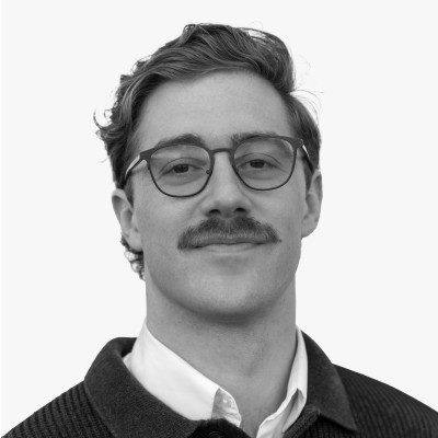 Head-and-shoulders portrait of Tim Robinson, wearing glasses, with a moustache, in a dark knit sweater over a white collar.